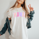 Ivory graphic tee with bold font that says mama in uppercase letters.  The fun summer design has bright purple, dark pink, and peach lettering.  Attached to the letters are white daisies.  