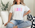 white graphic tee with bold font that says mama in uppercase letters.  The fun summer design has bright purple, dark pink, and peach lettering.  Attached to the letters are white daisies.  