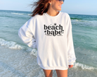 beach sweatshirt that says beach babe with small outlines or birds, sun, a palm tree, and wave lines.  Design is black and the sweatshirt is white.