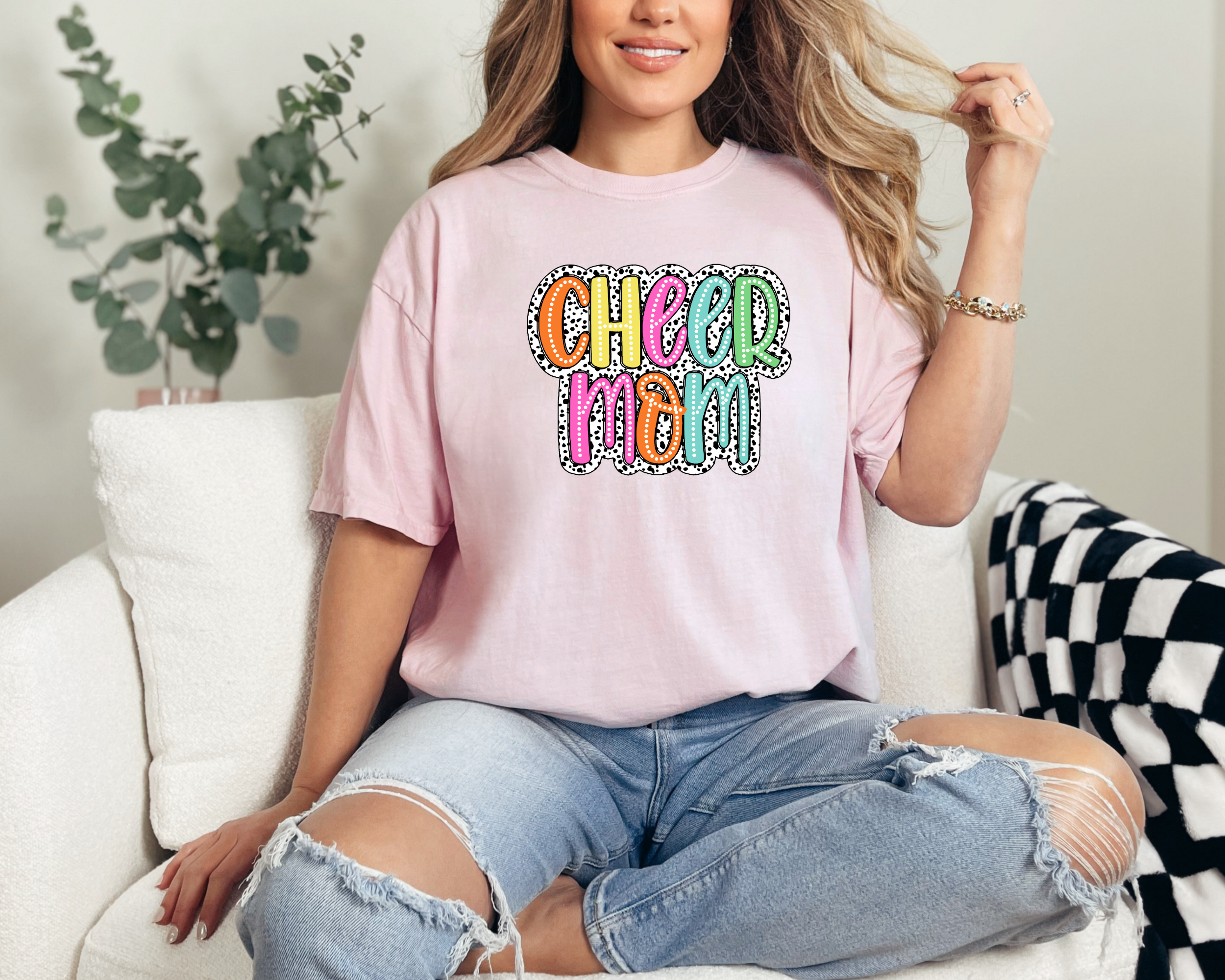 Cheer mom shirt in the color blossom with a bright doodle style font with a background behind the words that appears like black and white dalmatian dots.