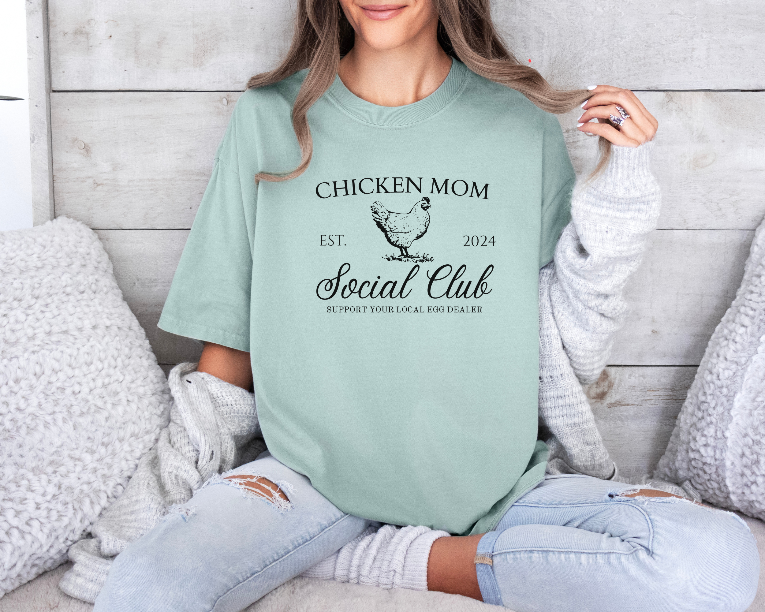 Funny chicken mom shirt that says chicken mom up top and in the middle a picture of a chicken.  On the left of the chicken it says est. and the right it says 2024.  Below that it says social club in fancy cursive and lastly below that it says support your local egg dealer.