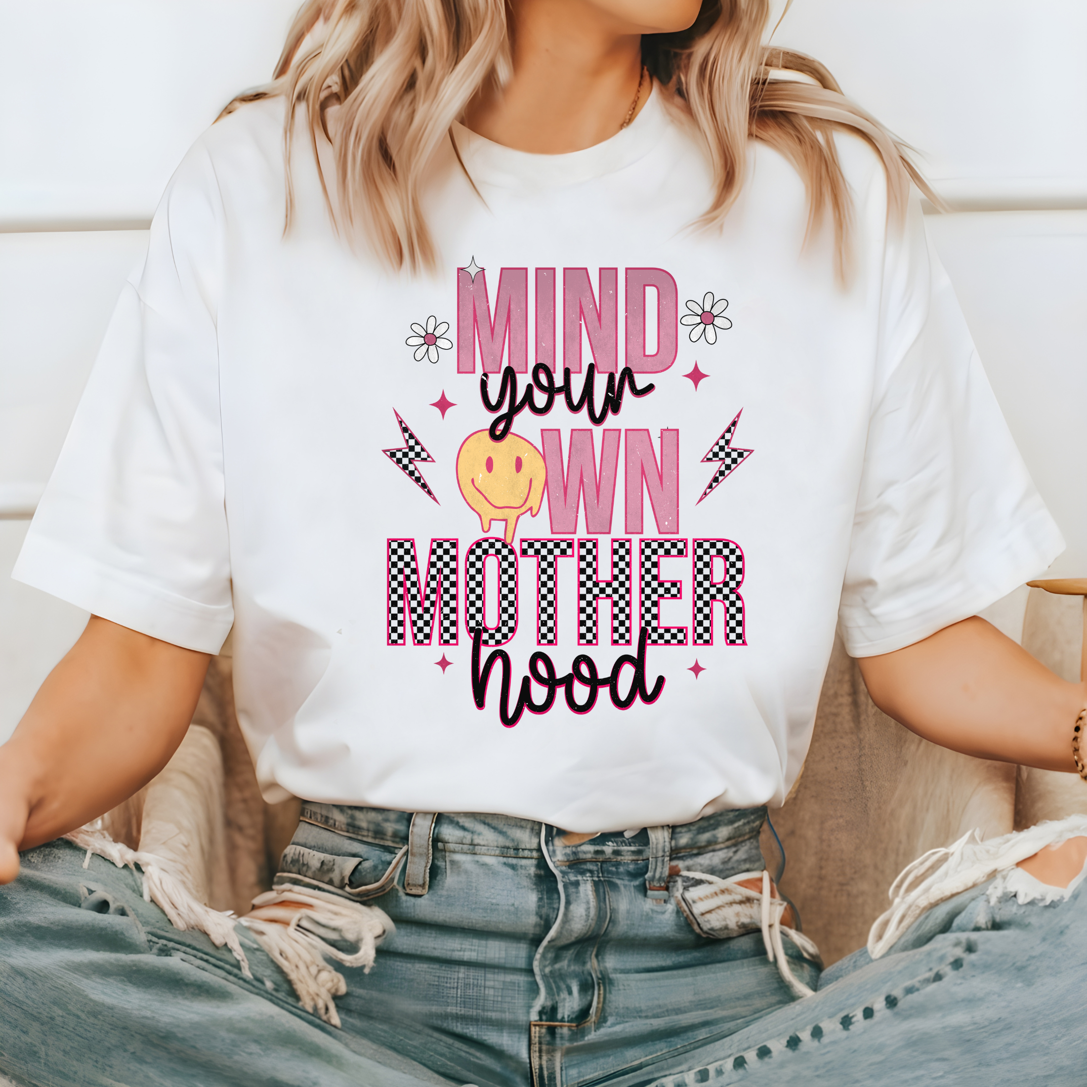 white mom graphic tee with a 90s theme design.  The design says mind your own motherhood in pink, checkered black and white and a yellow melting smiley face.  There are also two small daisies.  