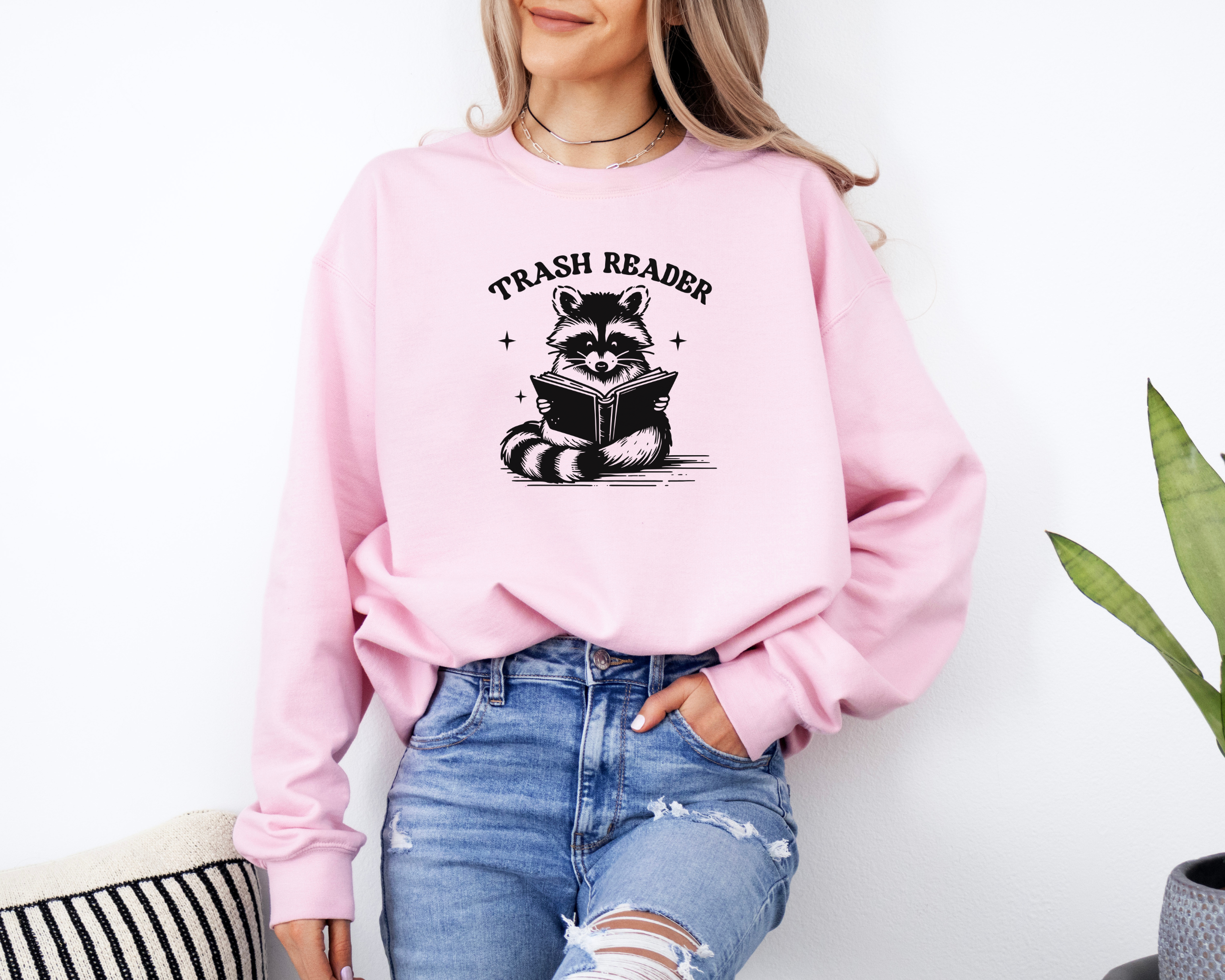 Trash reader with a raccoon reading a book.  The design is black and sweatshirt color is pink.